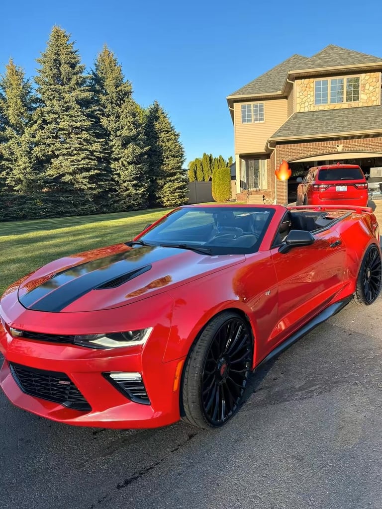 Red Chevrolet Camaro convertible parked in driveway with yellow two-story house and evergreen trees in background